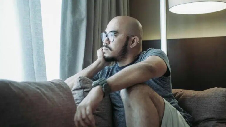 A bald man with a beard and glasses sits on a couch, looking pensively out a window.