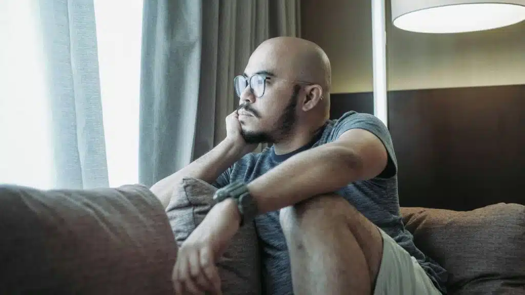 A bald man with a beard and glasses sits on a couch, looking pensively out a window.