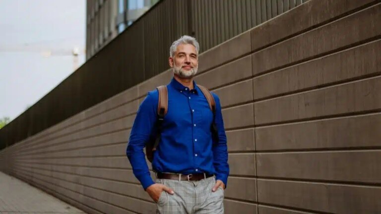 A stylish, gray-haired man in a blue shirt and backpack smiles outdoors.
