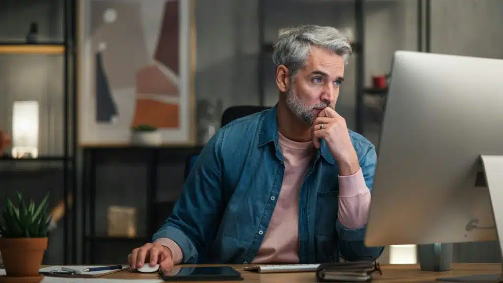 A focused man with gray hair and a beard uses a computer at a desk.