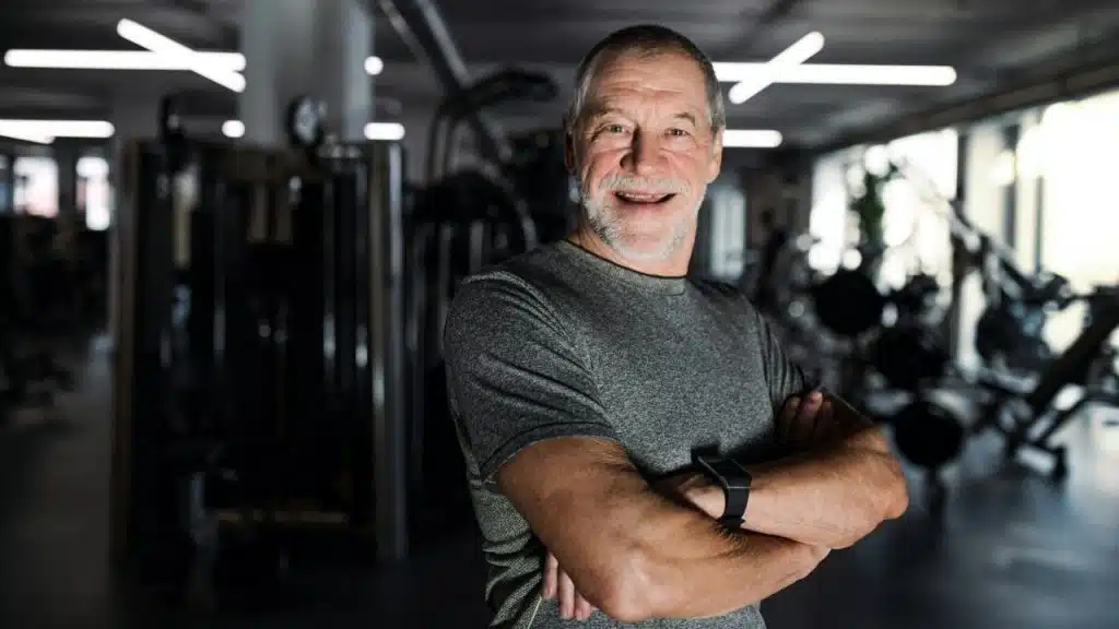 A smiling, bearded man with grey hair stands with arms crossed in a gym.