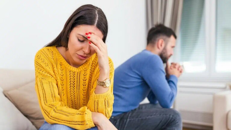 A distressed woman in a yellow sweater holds her head, while a man sits sadly in the background.