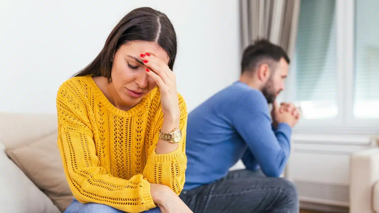 A distressed woman in a yellow sweater holds her head, while a man sits sadly in the background.