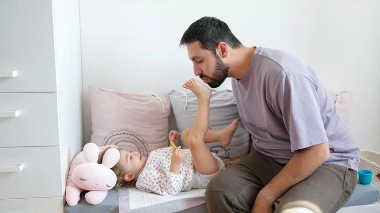 A man kissing a child’s foot as she lies on a cushion beside a stuffed bunny.