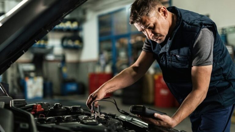 A mechanic in a gray shirt and dark blue vest works on a car engine.