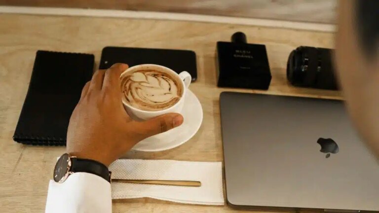 A person's hand reaches for a latte on a wooden table with a laptop and accessories.