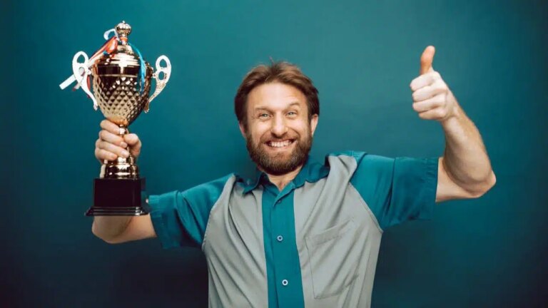 A bearded man in a two-tone shirt holds a trophy and gives a thumbs-up, smiling.