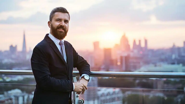 A smiling bearded man in a suit stands on a balcony overlooking a city.