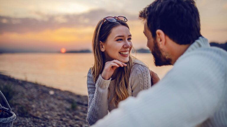 A couple smiling and talking by a lakeside at sunset.