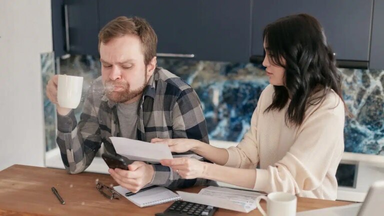 A man spits out a drink while looking at a phone, and a woman next to him holds papers.